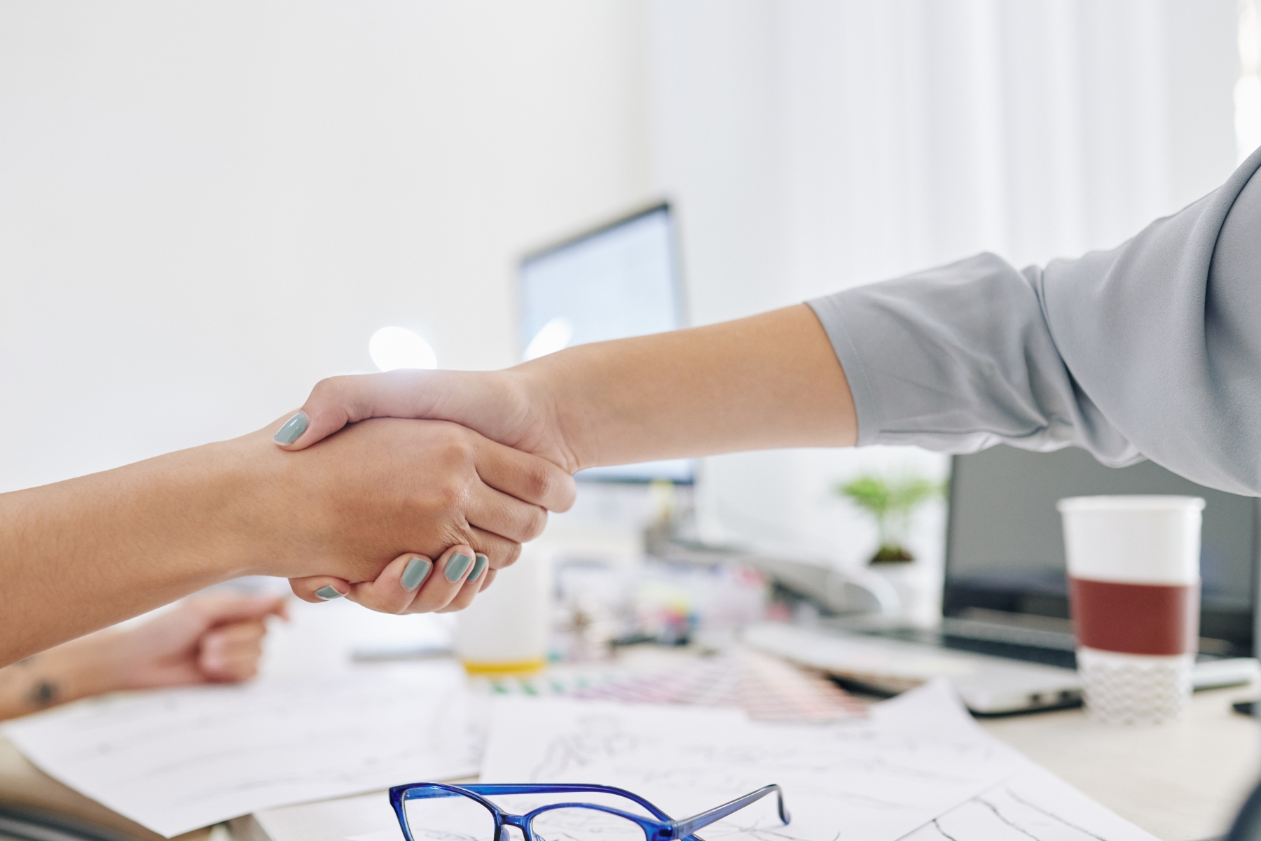 An image of two people shaking hands up close with a cluttered desk in the background